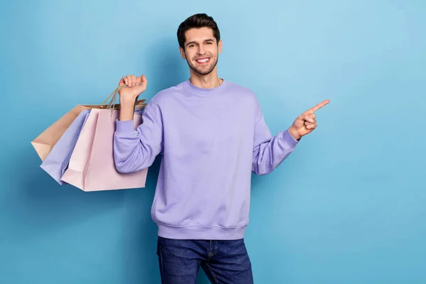 Photo of cute young brunet guy hold bags index promo wear sweater jeans isolated on blue color background — Foto de Stock