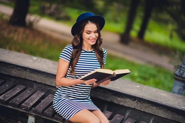 Portrait of attractive cheerful focused girl sitting on bench reading interesting academic book on fresh air outdoors — Stock Fotó