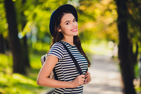 Фото очаровательной молодой леди носить полосатый рюкзак платья головной убор улыбаясь ходьба наслаждаясь хорошей погодой на открытом воздухе городской парк — стоковое фото