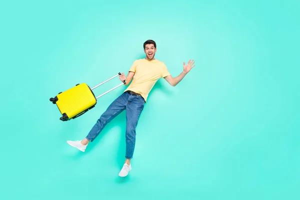Full body photo of funky brunet guy wave with bag wear t-shirt jeans sneakers isolated on teal background — Zdjęcie stockowe