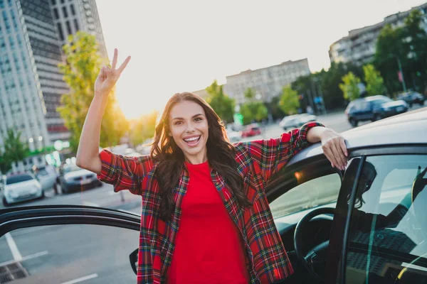 Retrato de atractiva chica alegre de pelo ondulado conductor mostrando v-signo de divertirse atardecer tiempo de la noche en el aparcamiento al aire libre —  Fotos de Stock
