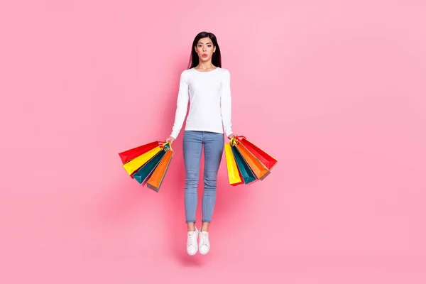 Full length photo of impressed young lady jump hold bags wear shirt jeans sneakers isolated on pink background — Stockfoto
