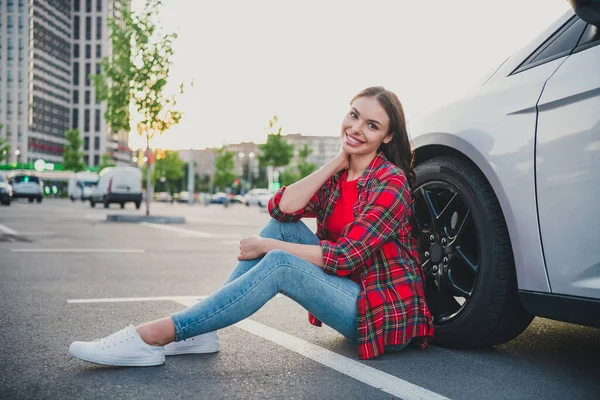 Portrait of attractive cheerful girl driver sitting on floor near new modern car ownership dealer on parking outdoors