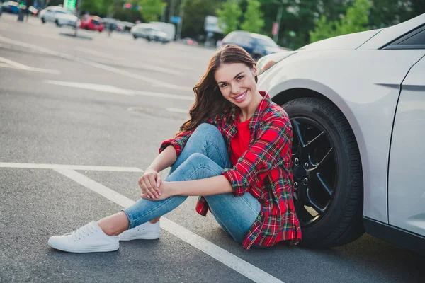 Retrato de atractiva chica alegre de pelo ondulado conductor nuevo propietario de coche sentado en el suelo descansando fin de semana buen humor en el aparcamiento al aire libre —  Fotos de Stock