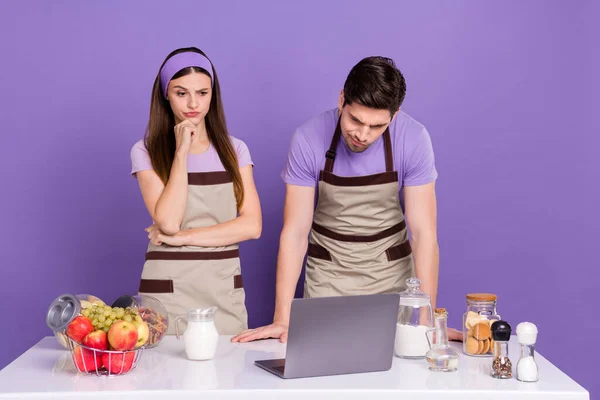 Photo of cute thoughtful boyfriend girlfriend dressed chef outfits reading recipes modern gadget isolated purple color background — Stockfoto