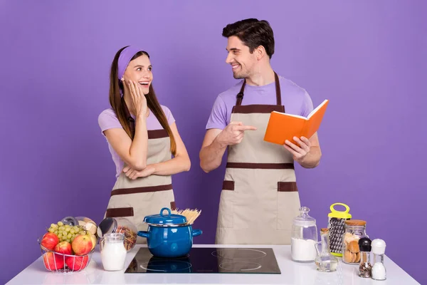 Foto de marido positivo divertido esposa usar delantales grises lectura apuntando libro de cocina aislado color violeta fondo — Foto de Stock