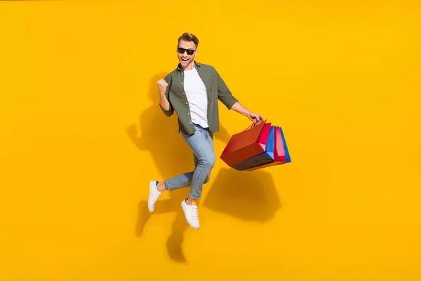 Volledige lichaamsomvang uitzicht van aantrekkelijke vrolijke man springen kopen geschenken dingen verheugen geïsoleerd over helder gele kleur achtergrond — Stockfoto