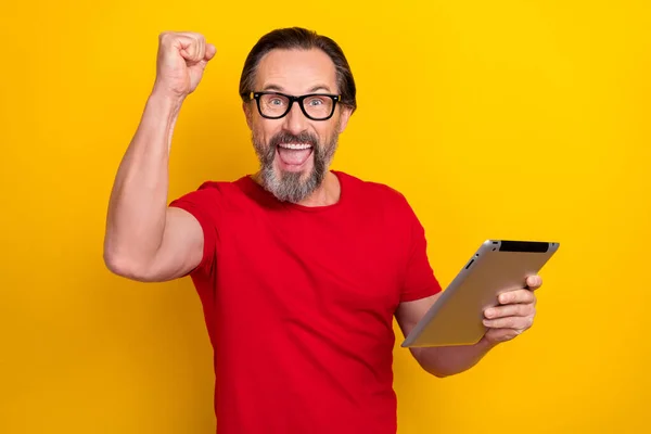 Photo of pretty lucky guy pensioner dressed red t-shirt glasses reading gadget rising fist isolated yellow color background — Stockfoto