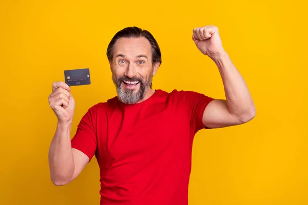 Photo of lucky funky retired man wear red t-shirt rising fist bank card isolated yellow color background — Foto de Stock
