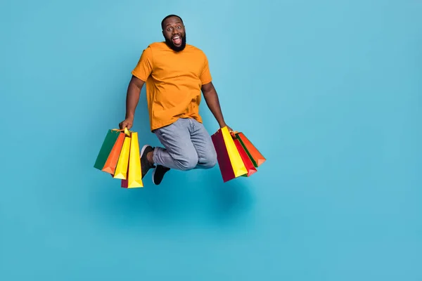 Photo of funky impressed guy dressed orange t-shirt jumping high holding bargains isolated blue color background — стоковое фото