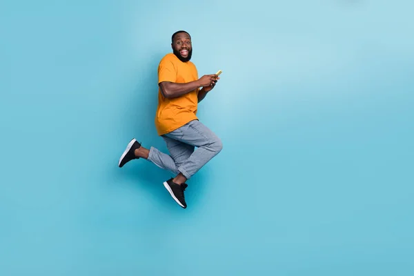Photo of excited cheerful guy dressed orange t-shirt jumping running chatting modern gadget isolated blue color background