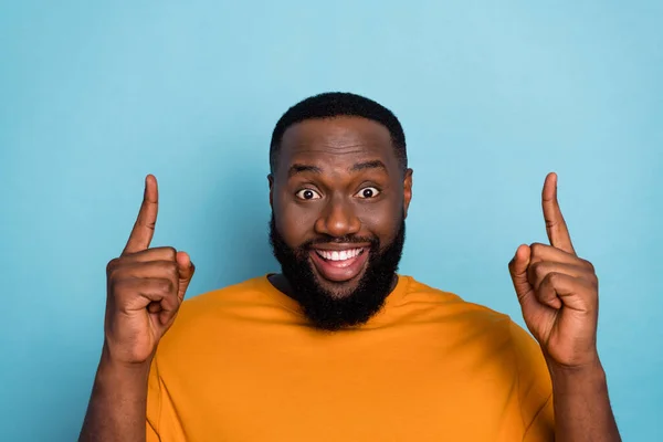 Photo of cute impressed guy wear orange t-shirt pointing up empty space isolated blue color background — стоковое фото