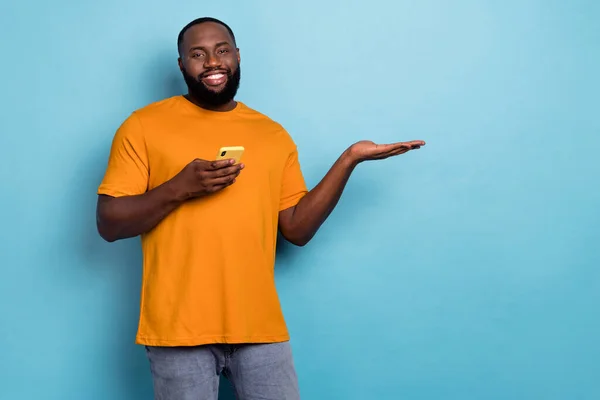 Photo of positive funky guy wear orange t-shirt chatting modern device holding arm empty space isolated blue color background — Photo