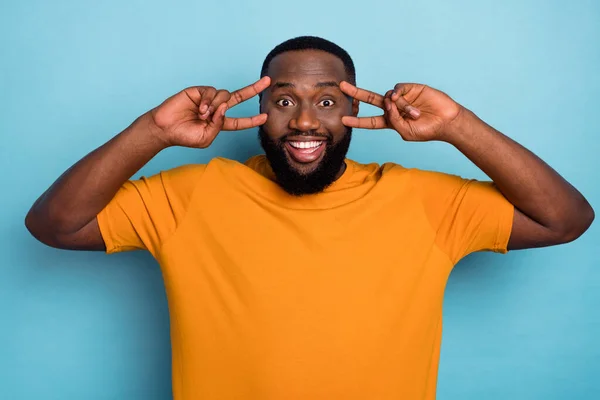 Photo of cheerful pretty guy wear orange t-shirt v-signs cover eyes isolated blue color background — Zdjęcie stockowe