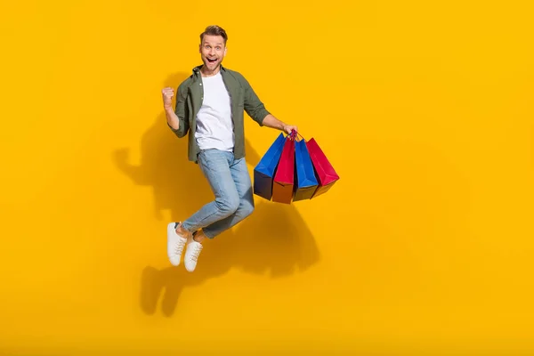 Повнометражний вигляд тіла привабливого щасливого веселого хлопця, який стрибає, купуючи подарунки, радіючи ізольованим на яскраво-жовтому кольоровому фоні — стокове фото