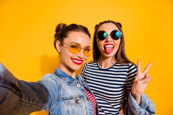 Портрет двох позитивних безтурботних дівчат, які беруть селфі, показують язик v-знак ізольовано на жовтому тлі — стокове фото