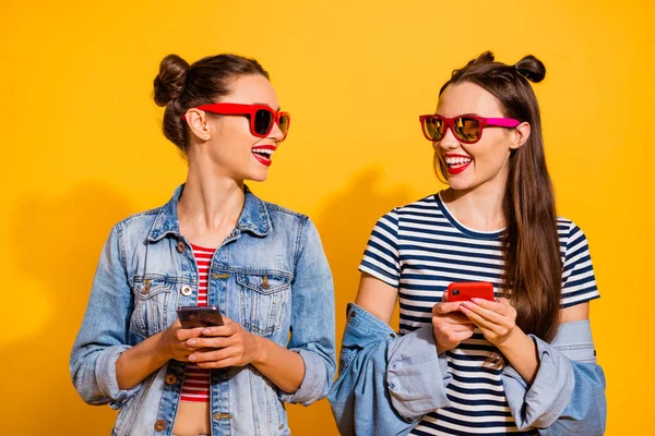 Portrait of two cheerful charming girls toothy smile hold telephone blogging isolated on yellow color background — Fotografia de Stock