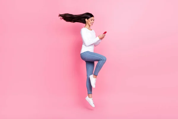 Full längd foto av söt tusenårig dam hoppa titta telefon bära skjorta jeans sneakers isolerad på rosa bakgrund — Stockfoto