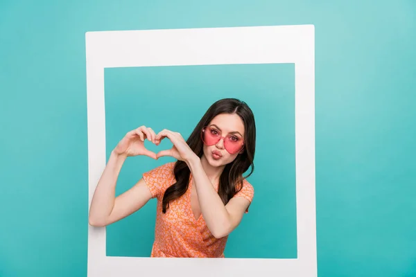 Foto de mulher doce brilhante usar roupa laranja óculos de pé dentro do quadro de papel mostrando coração isolado cor turquesa fundo — Fotografia de Stock