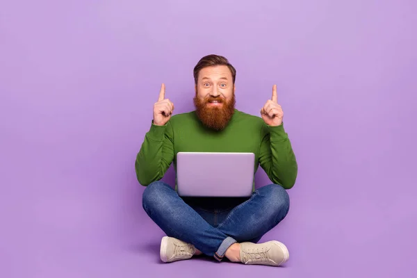 Full body portrait of positive glad man sit floor indicate fingers up empty space advert isolated on purple color background