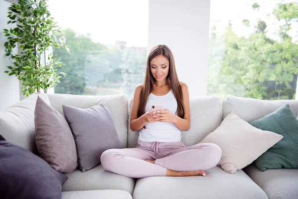 Full size photo of pretty positive young lady sitting sofa use hold phone chatting write typing blog comment indoors — Stock fotografie