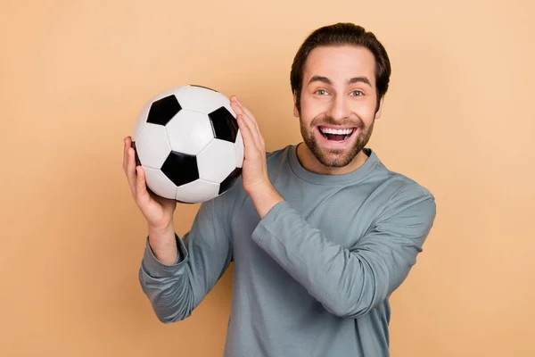 Portrét spokojený rád muž paže držet fotbal zubatý úsměv vzhled fotoaparát izolované na béžové barvy pozadí — Stock fotografie