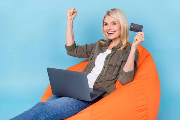 Excited aged lady sit bag chair use netbook buy purchase credit card get jackpot bet fist up isolated blue color background — Stock Photo, Image