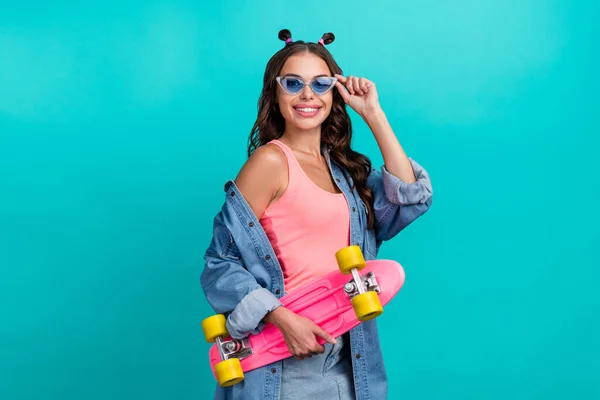 Foto van jong vrolijk mooi meisje houden skateboard sportieve brillen geïsoleerd over turquoise kleur achtergrond — Stockfoto