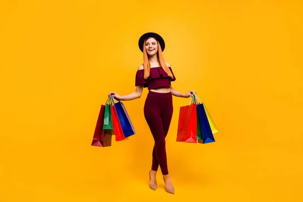 Teljes hosszúságú fotó aranyos izgatott hölgy öltözött piros ruhák fejfedő gazdaság vásárlók üres tér elszigetelt sárga szín háttér — Stock Fotó