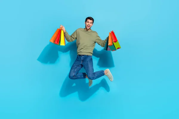 Volledige foto van koele duizendjarige brunet jongen springen met tassen dragen shirt jeans schoenen geïsoleerd op blauwe achtergrond — Stockfoto