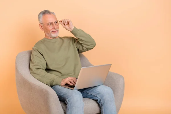 Foto de otimista envelhecido cinza penteado homem sentar trabalho laptop olhar promo desgaste óculos pulôver jeans isolado no fundo bege — Fotografia de Stock