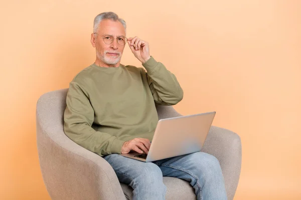 Zdjęcie surowego starszego siwego fryzjera siedzieć praca laptop nosić okulary pullover dżinsy izolowane na beżowym tle — Zdjęcie stockowe