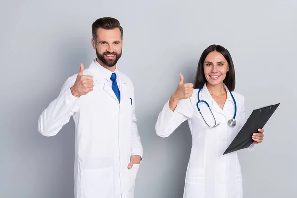 Foto de dos docs sonrientes inteligentes vestidos con batas blancas sujetando los pulgares del portapapeles hacia arriba fondo gris aislado — Foto de Stock