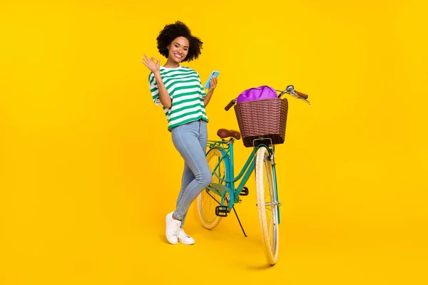 Foto de comprimento total de doce jovem senhora perto de bicicleta segurar telefone okey desgaste t-shirt jeans sapatilhas saco isolado no fundo amarelo — Fotografia de Stock
