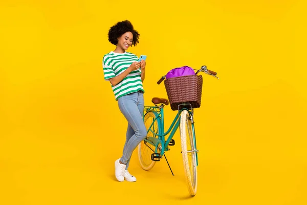 Foto em tamanho completo de senhora millennial bonito perto de bicicleta olhar telefone desgaste t-shirt jeans sapatos saco isolado no fundo amarelo — Fotografia de Stock
