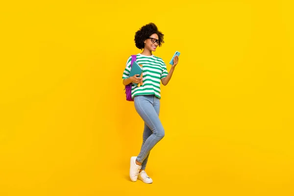 Full body foto van schattige jonge dame kijken telefoon hold boek dragen bril t-shirt tas jeans sneakers geïsoleerd op gele achtergrond — Stockfoto
