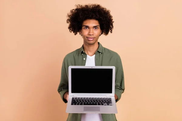 Портрет привабливого серйозного модного хвилястого хлопця, який тримає рекламу ноутбука, ізольовану на бежевому пастельному кольоровому фоні — стокове фото
