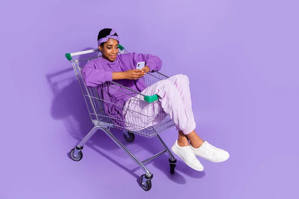 Foto completa de trans bisexual hipster masculino sit shopping cart surfeando en el teléfono aislado sobre fondo de color violeta — Foto de Stock