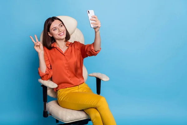 Çekici, neşeli bir kızın portresi ofis koltuğunda oturmuş V-sgn 'un parlak mavi arka planda izole edilmiş selfie çekişini gösteriyor. — Stok fotoğraf