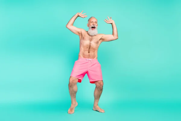 Photo of carefree old man dance beach party rejoice raise arms wear pink shorts barefoot isolated teal color background.
