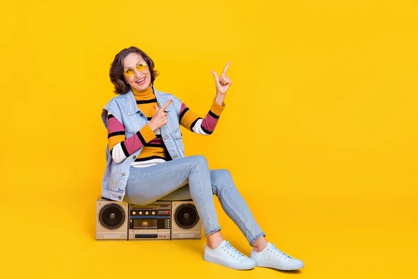Full body photo of curious grey hairdo elder lady sit on boom box index wear eyewear sweater vest jeans shoes isolated on yellow background.