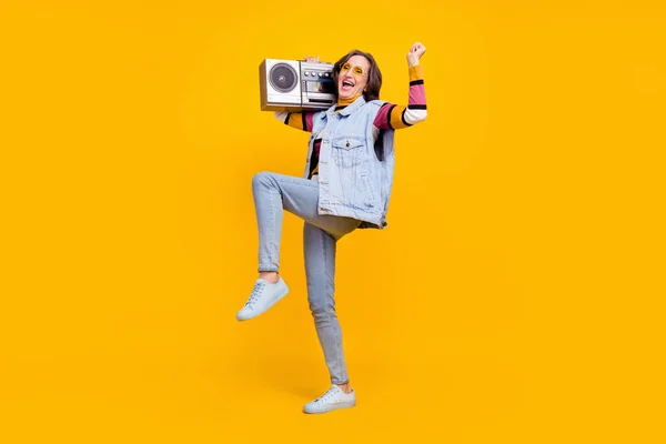Full length photo of optimistic white hairdo elder lady hold boom box wear eyewear sweater vest jeans shoes isolated on yellow color background.