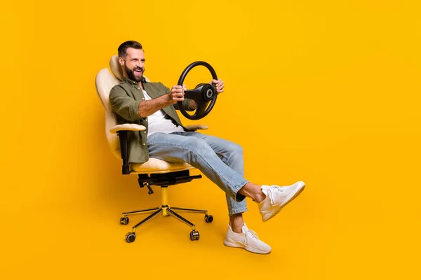 Full body profile side photo of young guy have fun play videogame steering wheel sit stool isolated over yellow color background.