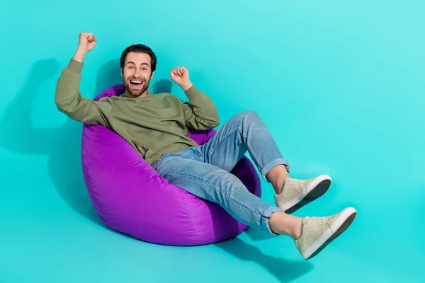 Full body profile side photo of young man sit purple chair rejoice victory fists hands isolated over turquoise color background.