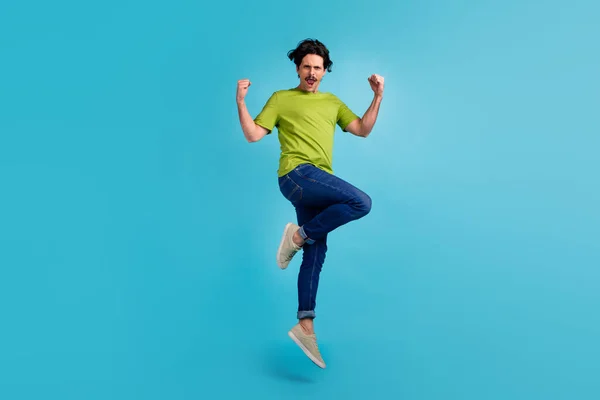 Full length photo of funky lucky guy dressed green t-shirt jumping high rising fists isolated blue color background.