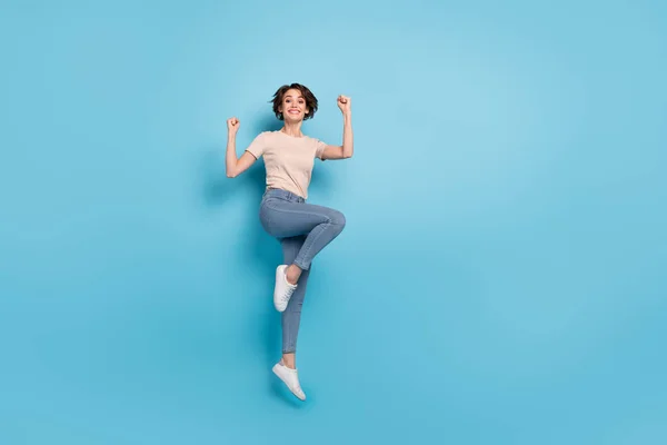 Full length photo of pretty lucky woman wear beige t-shirt jumping high rising fists empty space isolated blue color background.