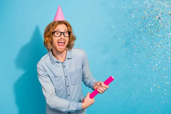 Photo of sweet shocked young guy dressed jeans shirt eyewear headwear shooting confetti isolated blue color background.