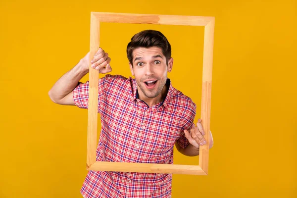 Photo of cheerful young amazed man hold hands wooden frame creative isolated on yellow color background.