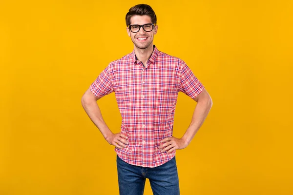 Photo of positive happy young cheerful man hold hands waist smile wear glasses isolated on yellow color background.