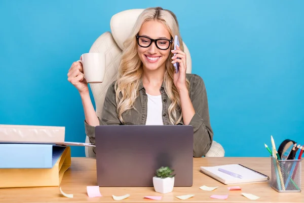 照片肖像女士坐在办公室的笔记本电脑上 一边喝着咖啡一边用手机聊着孤立的淡蓝色背景 — 图库照片